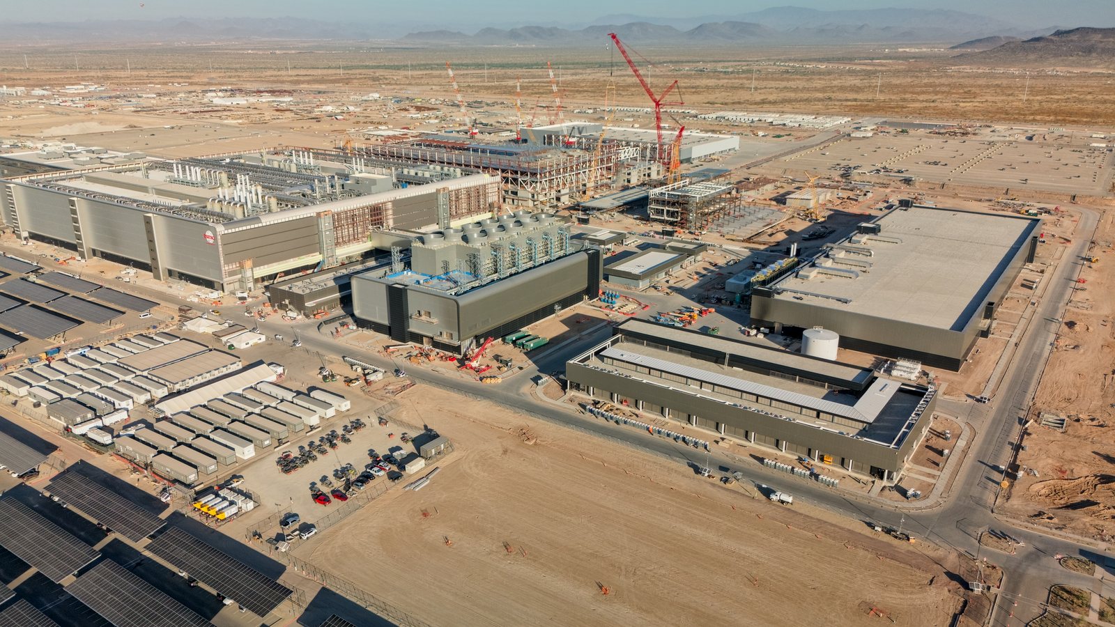 Aerial view of a vast industrial site under construction in the desert, with cranes, grey buildings and car parks covered with solar panels