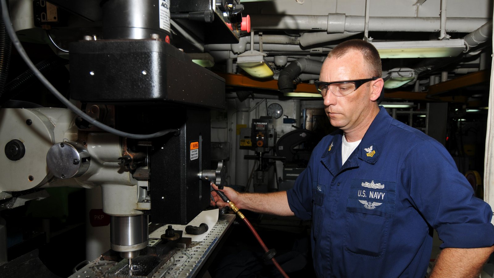 A sailor in a blue US Navy uniform and safety glasses stands at a machine tool in a workshop with exposed pipework